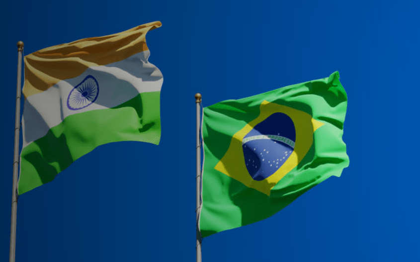 The national flags of India and Brazil fly side by side on flagpoles against a clear blue sky, their colors visible as the fabric moves in the wind.