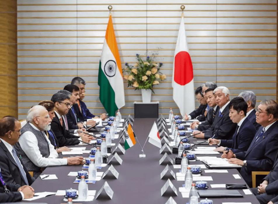 Delegations from India and Japan sit facing each other at a long conference table during a formal bilateral meeting, with national flags displayed behind them, documents and microphones arranged neatly as officials engage in diplomatic discussion.