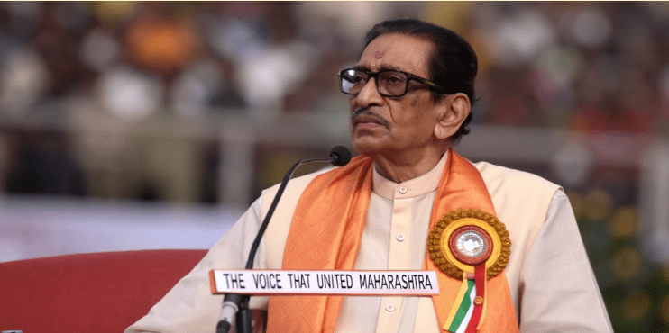 Balasaheb Thackeray sits on a stage wearing an orange shawl and glasses, speaking into a microphone during a public event, with a badge on his chest and a blurred crowd in the background, symbolizing his role as a prominent voice in Maharashtra politics.