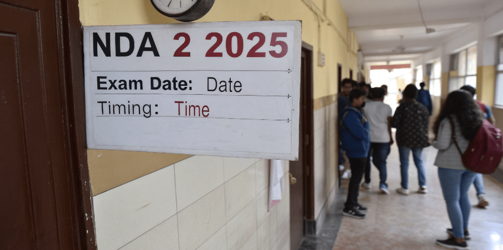 NDA 2 2025 exam date and timing notice board in school corridor with students, showing shift schedule for Mathematics and GAT papers.