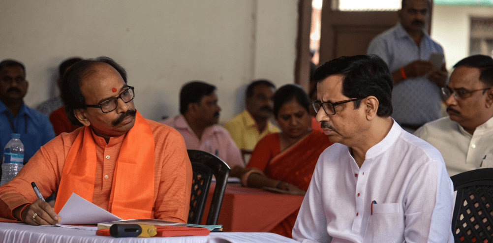 Two senior Indian political leaders seated at a public meeting, one in an orange scarf writing notes on paper while the other in a white kurta listens attentively, with party members seated behind them in an indoor hall.