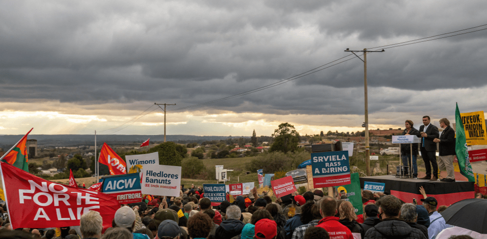 Large outdoor political rally with a dense crowd holding protest and party banners, speakers standing on a raised stage with microphones, rural landscape and houses in the background, and dark storm clouds overhead, symbolizing a major coalition-era political gathering.